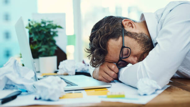 Na slici je prikazan muškarac koji je zaspao za stolom usled gomile obaveza i stresa na poslu - odnosi se na to kako stres sabotira ravan stomak kod muškaraca 40+