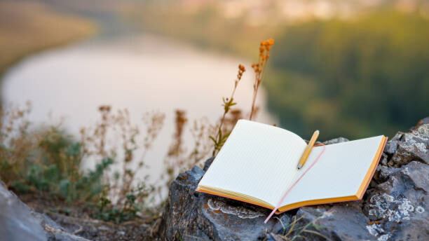 Na slici je prikazana sveska i olovka, okružene prirodom - odnosi se na važnost samorefleksije.